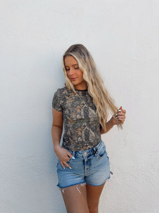 Person wearing a camouflage shirt and denim shorts against a white wall