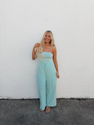 Woman in an aqua jumpsuit standing by a white wall with a gray floor.