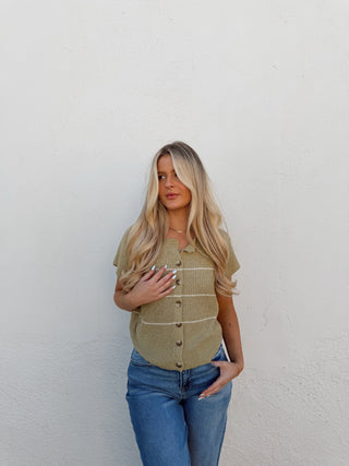 Woman wearing a green vest and blue jeans against a white wall