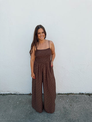 Woman in a brown polka dot jumper standing by  a white wall and gray pavement.