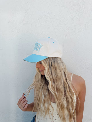 Person with long blonde hair wearing a white cap with a blue visor on a plain background