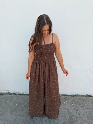 Woman in a brown polka dot jumper standing by  a white wall and gray pavement.